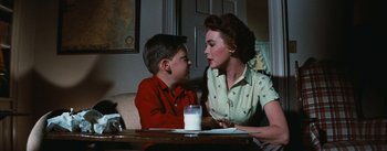 Movie still from “Bigger Than Life” (1956), directed by Nicholas Ray – A woman and a boy sitting at a table with a glass of milk in front of them; Medium shot, Over the shoulder angle