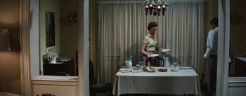 Movie still from “Bigger Than Life” (1956), directed by Nicholas Ray – A woman standing in front of a table with plates of food on top of it; Wide shot, High angle