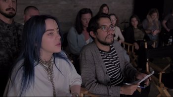 Movie still from “Billie Eilish: The World's a Little Blurry” (2021), directed by R.J. Cutler – A man and a woman sitting in front of other people; Close Up shot, High angle