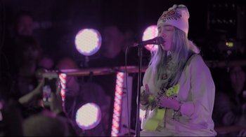 Movie still from “Billie Eilish: The World's a Little Blurry” (2021), directed by R.J. Cutler – A woman in a white shirt is playing a guitar; Medium shot, Low angle