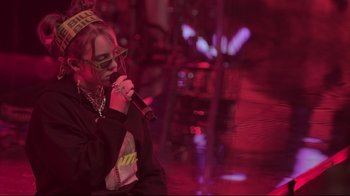 Movie still from “Billie Eilish: The World's a Little Blurry” (2021), directed by R.J. Cutler – A person with a microphone in front of a red background; Medium shot, Over the shoulder angle