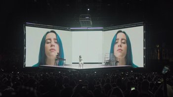 Movie still from “Billie Eilish: The World's a Little Blurry” (2021), directed by R.J. Cutler – A woman standing on a stage in front of a large screen; Extreme Wide shot, Low angle