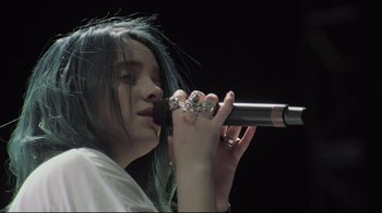 Movie still from “Billie Eilish: The World's a Little Blurry” (2021), directed by R.J. Cutler – A woman with blue hair is holding a microphone; Close Up shot, Low angle