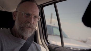 Movie still from “Billie Eilish: The World's a Little Blurry” (2021), directed by R.J. Cutler – An old man with a beard and glasses sitting in a car; Close Up shot, Over the shoulder angle