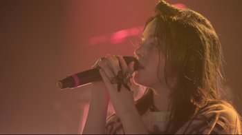 Movie still from “Billie Eilish: The World's a Little Blurry” (2021), directed by R.J. Cutler – A woman is singing into a microphone on a stage; Close Up shot, Low angle