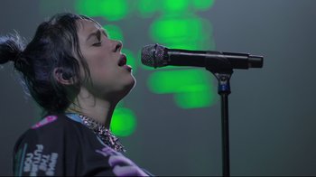 Movie still from “Billie Eilish: The World's a Little Blurry” (2021), directed by R.J. Cutler – A woman singing into a microphone on a stage; Close Up shot, Low angle