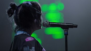 Movie still from “Billie Eilish: The World's a Little Blurry” (2021), directed by R.J. Cutler – A person with a microphone on a stage; Close Up shot, Low angle