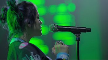 Movie still from “Billie Eilish: The World's a Little Blurry” (2021), directed by R.J. Cutler – A woman is holding a microphone up to a microphone stand; Close Up shot, Low angle