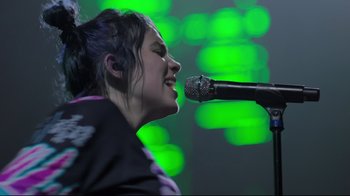 Movie still from “Billie Eilish: The World's a Little Blurry” (2021), directed by R.J. Cutler – A woman singing into a microphone on a stage; Close Up shot, Low angle