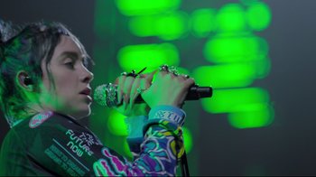 Movie still from “Billie Eilish: The World's a Little Blurry” (2021), directed by R.J. Cutler – A person holding a microphone on a stage; Medium shot, Low angle