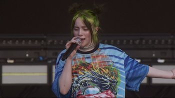 Movie still from “Billie Eilish: The World's a Little Blurry” (2021), directed by R.J. Cutler – A woman with green hair is singing into a microphone; Medium shot, Low angle