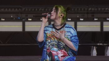 Movie still from “Billie Eilish: The World's a Little Blurry” (2021), directed by R.J. Cutler – A woman with green hair is singing into a microphone; Medium shot, High angle