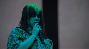 Movie still from “Billie Eilish: The World's a Little Blurry” (2021), directed by R.J. Cutler – A woman with green hair is holding a microphone; Medium shot, Low angle