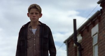Movie still from “Billy Elliot” (2000), directed by Stephen Daldry – A young boy standing in front of a brick building; Medium shot, Low angle