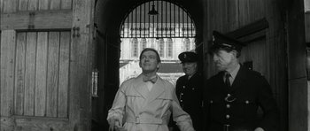 Movie still from “Billy Liar” (1963), directed by John Schlesinger – A man in a trench coat and a tie; Medium shot, Low angle