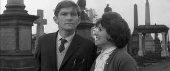 Movie still from “Billy Liar” (1963), directed by John Schlesinger – A man and a woman standing next to each other in front of a statue; Medium shot, Low angle