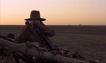 Movie still from “Billy Two Hats” (1974), directed by Ted Kotcheff – A man sitting on top of a log in the middle of a field taking a picture; Wide shot, Low angle