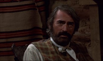 Movie still from “Billy Two Hats” (1974), directed by Ted Kotcheff – A person sitting in a chair wearing a vest; Close Up shot, Over the shoulder angle