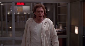 Movie still from “Bio-Dome” (1996), directed by Jason Bloom – A man with long hair and a white t - shirt is standing in front of a counter; Medium shot, Low angle