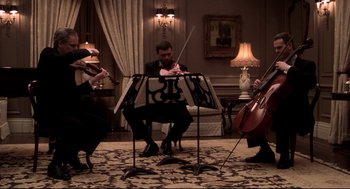 Movie still from “Birth” (2004), directed by Jonathan Glazer – A man sitting at a table playing a violin while two people play the cello; Wide shot, Low angle