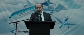 Movie still from “Black Box” (2021), directed by Yann Gozlan – A man in a black suit is speaking into a microphone; Close Up shot, Low angle