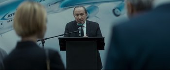 Movie still from “Black Box” (2021), directed by Yann Gozlan – A man standing at a podium speaking into a microphone; Close Up shot, Over the shoulder angle