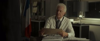 Movie still from “Black Box” (2021), directed by Yann Gozlan – An older man sitting at a desk looking at a piece of paper; Close Up shot, Over the shoulder angle