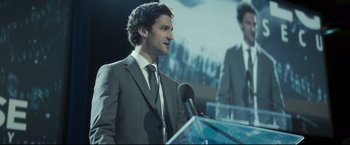 Movie still from “Black Box” (2021), directed by Yann Gozlan – A man standing at a podium speaking to a crowd; Medium shot, Over the shoulder angle