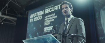 Movie still from “Black Box” (2021), directed by Yann Gozlan – A man standing at a podium in front of a projection screen; Medium shot, Over the shoulder angle