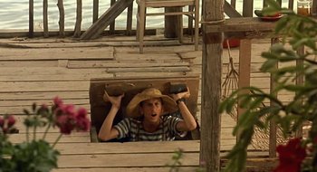 Movie still from “Black Cat, White Cat” (1998), directed by Emir Kusturica – A young man wearing a straw hat is peeking out of a window; Wide shot, High angle