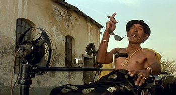 Movie still from “Black Cat, White Cat” (1998), directed by Emir Kusturica – An older man sitting in front of an old building; Medium shot, Low angle