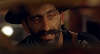 Movie still from “Black Cat, White Cat” (1998), directed by Emir Kusturica – A person wearing a hat with a mustache; Close Up shot, Over the shoulder angle