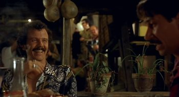 Movie still from “Black Cat, White Cat” (1998), directed by Emir Kusturica – A man sitting at a table in front of other people; Close Up shot, Over the shoulder angle