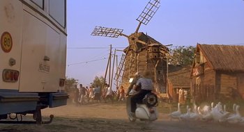 Movie still from “Black Cat, White Cat” (1998), directed by Emir Kusturica – A man riding a motorcycle in front of a windmill; Wide shot, Low angle