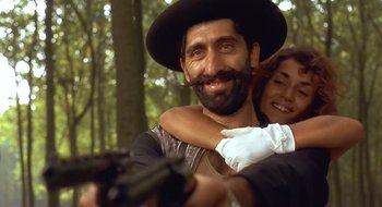 Movie still from “Black Cat, White Cat” (1998), directed by Emir Kusturica – A man with a beard hugging a woman with a gun; Close Up shot, Low angle