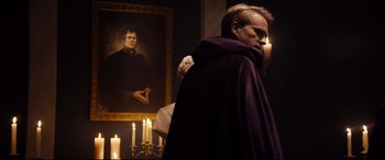 Movie still from “Black Christmas” (2019), directed by Sophia Takal – A man wearing a purple robe standing in front of a painting; Medium shot, Over the shoulder angle