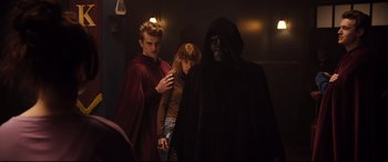 Movie still from “Black Christmas” (2019), directed by Sophia Takal – A man and a woman standing in front of a person in a hooded cloak; Medium shot, Over the shoulder angle