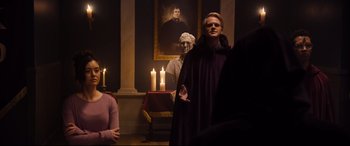 Movie still from “Black Christmas” (2019), directed by Sophia Takal – A man in a purple robe standing in front of a painting; Medium shot, Over the shoulder angle