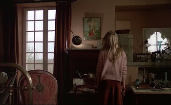Movie still from “Black Moon” (1975), directed by Louis Malle – A woman standing in front of a fireplace in a living room; Wide shot, High angle