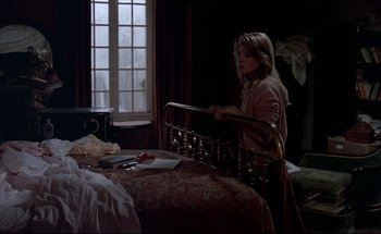 Movie still from “Black Moon” (1975), directed by Louis Malle – A woman sitting in a chair next to a bed in a room; Medium shot, Over the shoulder angle