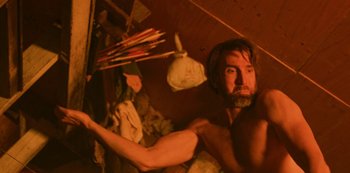 Movie still from “Ted K” (2021), directed by Tony Stone – A man with a beard and no shirt in a room with a bunch of stuff; Medium shot, Low angle