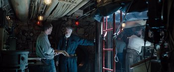 Movie still from “Black Sea” (2014), directed by Kevin Macdonald – A man standing next to another man in a room; Medium shot, Overhead angle