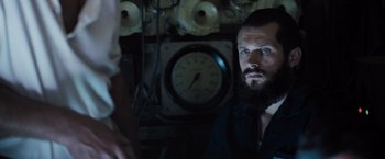 Movie still from “Black Sea” (2014), directed by Kevin Macdonald – A man with a long beard is sitting in front of a clock; Close Up shot, Over the shoulder angle