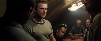 Movie still from “Black Sea” (2014), directed by Kevin Macdonald – A man standing next to another man in a dark room; Medium shot, Over the shoulder angle
