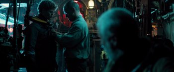 Movie still from “Black Sea” (2014), directed by Kevin Macdonald – A man standing in front of other people in a room; Medium shot, Over the shoulder angle