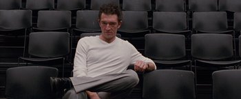 Movie still from “Black Swan” (2010), directed by Darren Aronofsky – A man sitting on a chair holding a newspaper in front of a row of black chairs; Medium shot, Low angle