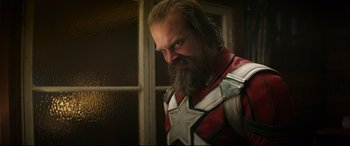 Movie still from “Black Widow” (2021), directed by Cate Shortland – A man with long hair and a beard wearing a red and white suit; Close Up shot, Over the shoulder angle