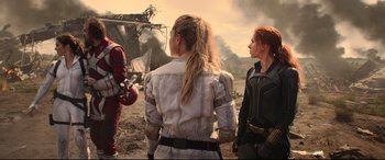 Movie still from “Black Widow” (2021), directed by Cate Shortland – A group of people standing next to each other on a dirt field; Medium shot, Over the shoulder angle