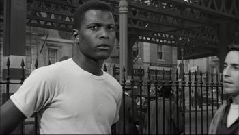 Movie still from “Blackboard Jungle” (1955), directed by Richard Brooks – A black and white photo of a man standing in front of an iron fence; Medium shot, Low angle