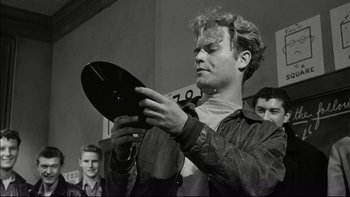 Movie still from “Blackboard Jungle” (1955), directed by Richard Brooks – A young man holding a record in front of a group of people; Close Up shot, Over the shoulder angle